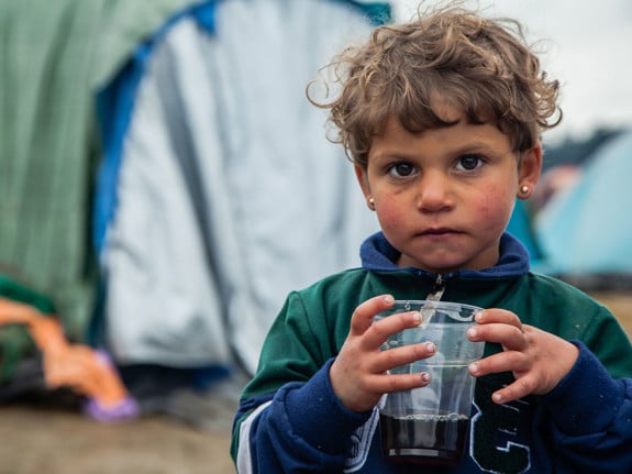 En liten flicka som håller i ett glas med te. I bakgrunden syns tält då hon befinner sig i ett flyktingläger.